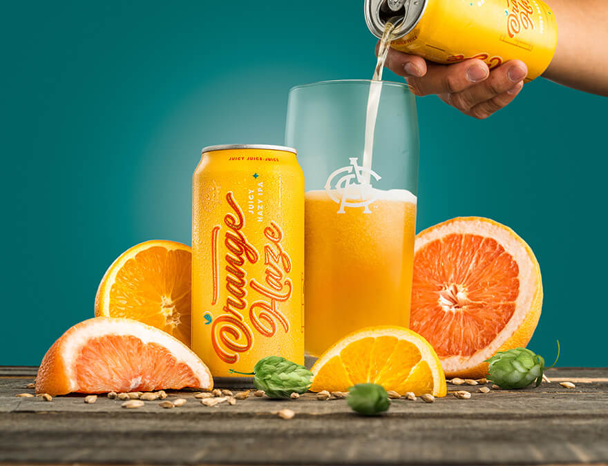 orange Haze can, and hand pouring juice over glass on a table with orange chunks
